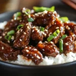 A close-up of a bowl of Spicy Firecracker Beef served over rice, garnished with green onions, sesame seeds, and extra sriracha.